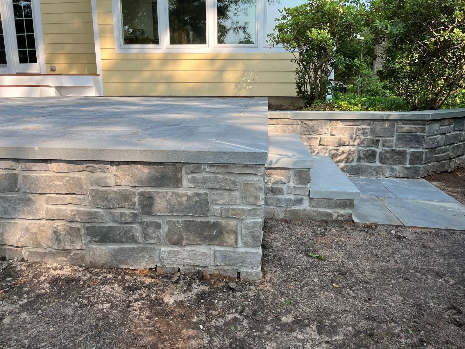 A stone patio and retaining wall with a single step leading down to a dirt ground next to a yellow-sided house.