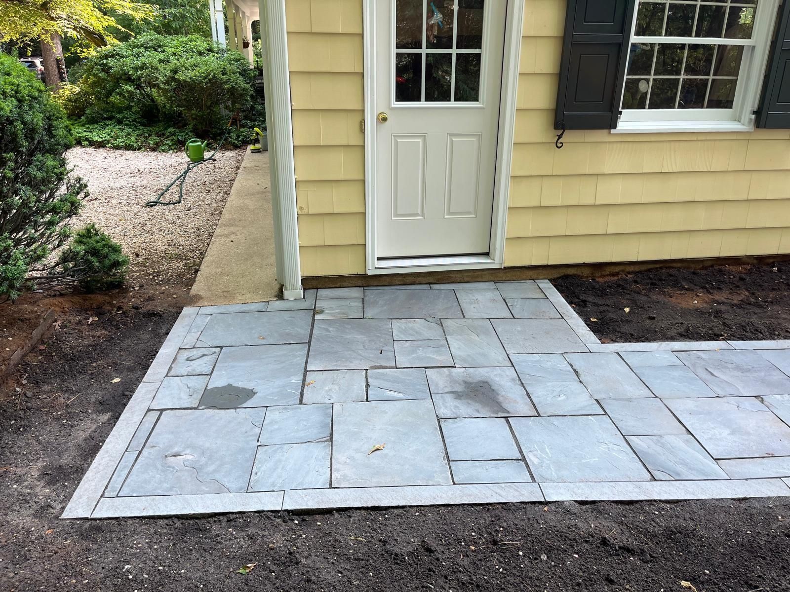 A rectangular flagstone patio leads to a light yellow house with a white door and dark shutters.