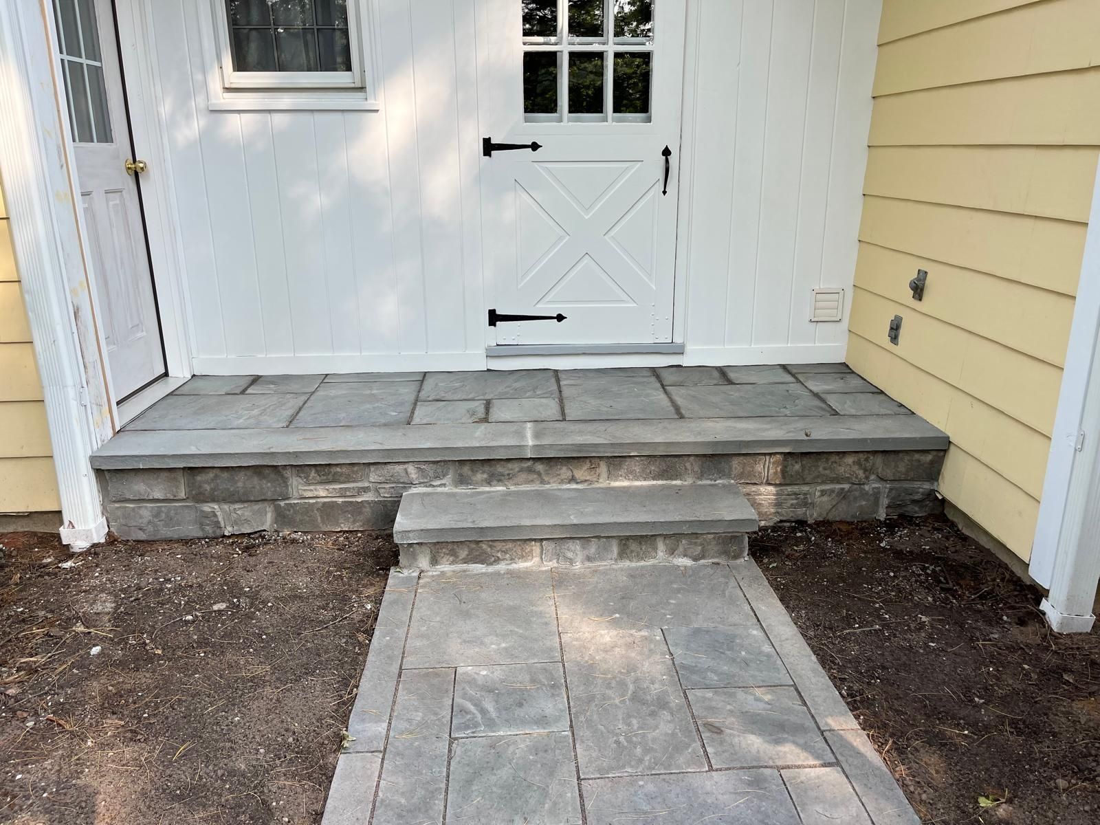 A gray stone patio and walkway lead to a white house door, featuring a single stone step at the entrance.