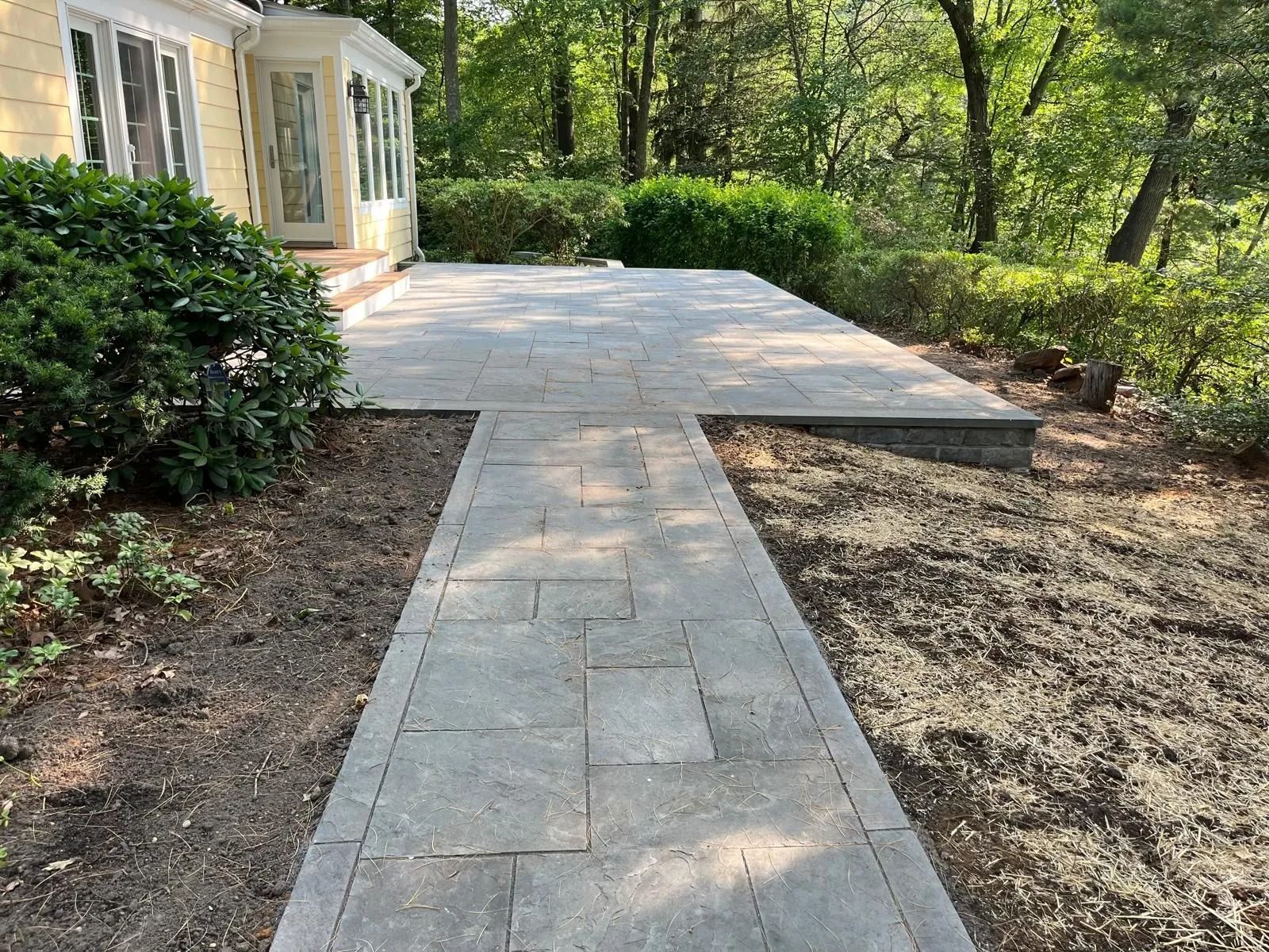 A stone walkway leads from the side of a yellow house to a paved patio in a wooded backyard.