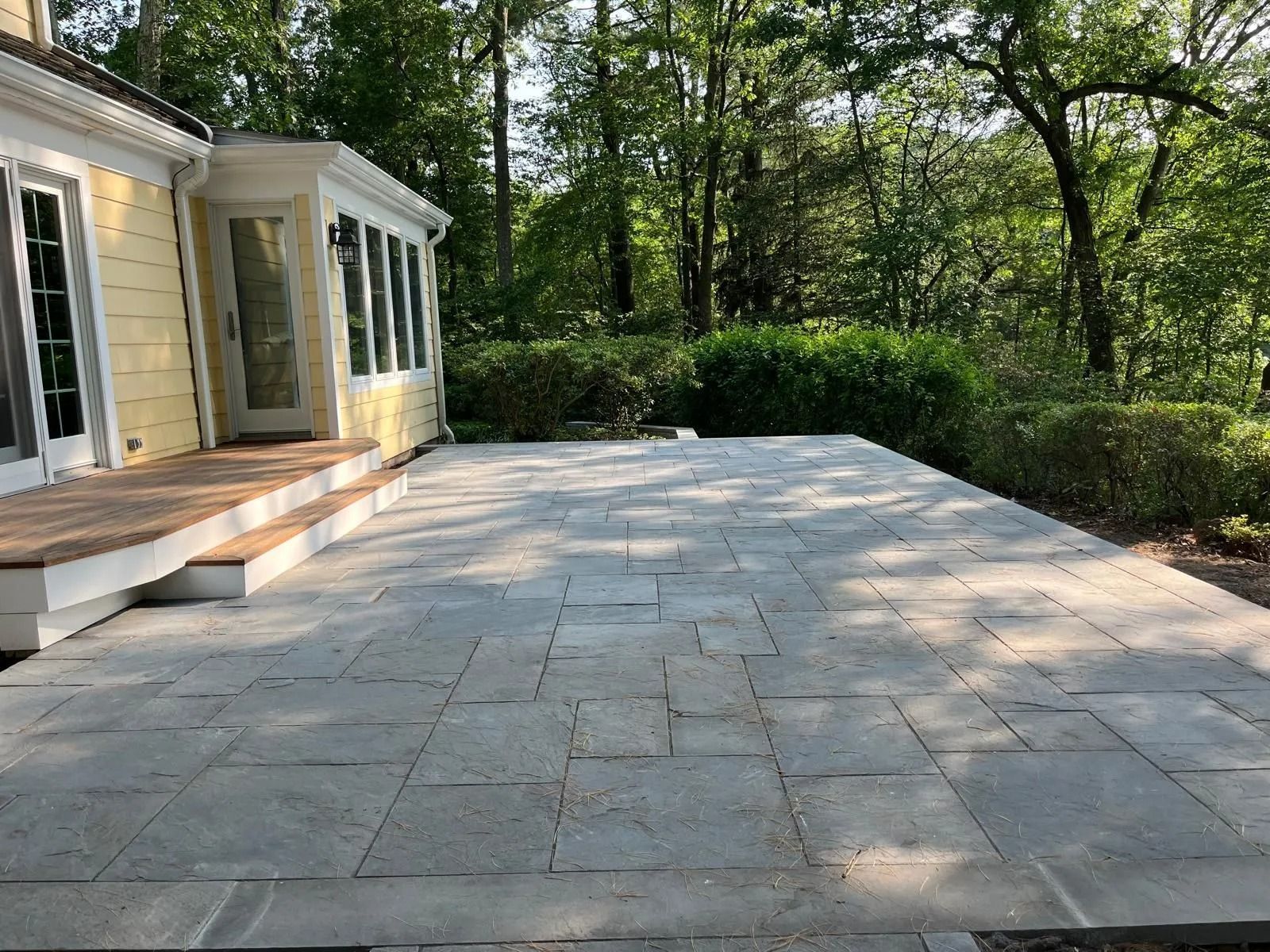 A wide, gray stone patio adjoins the wooden steps of a yellow house, surrounded by lush green trees and landscaping.