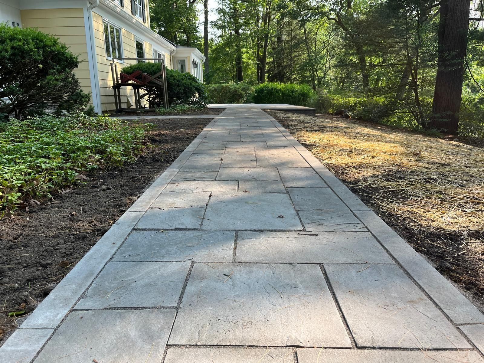 A paved walkway of rectangular grey stones leads toward a yellow house surrounded by trees and greenery.