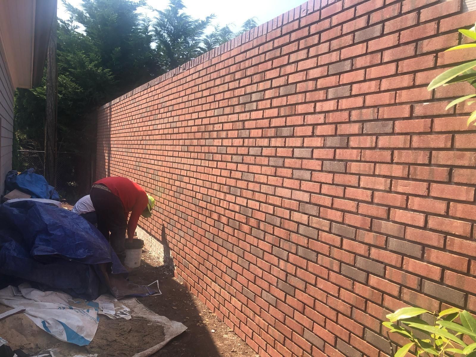 A bricklayer works on a long, red brick wall outdoors, with a pile of blue tarps and supplies beside them.