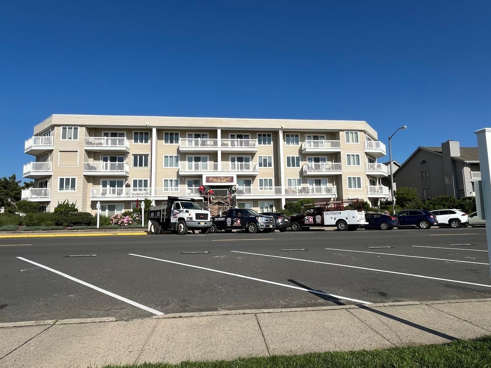 A multi-story beige apartment building with balconies stands behind a parking lot with utility trucks under a blue sky.