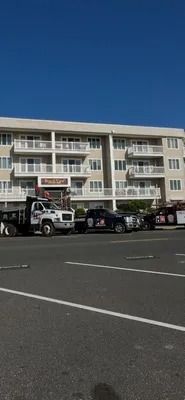 A large, light-colored apartment building with balconies, seen from a parking lot with service vehicles parked in front.