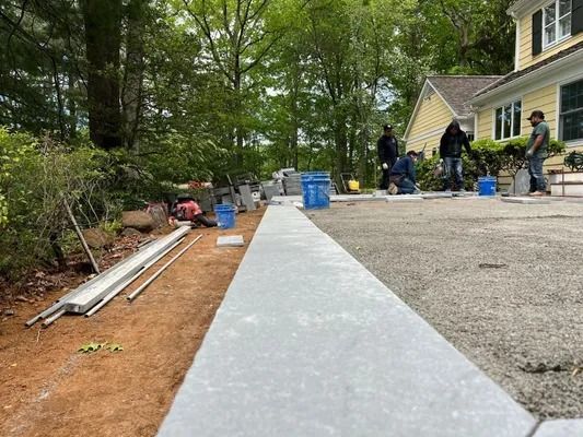 Workers build a stone patio next to a yellow house in a wooded yard.