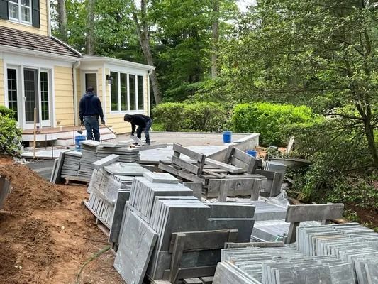 Two workers install stone pavers on a patio outside a yellow house, surrounded by stacked stone tiles and soil.