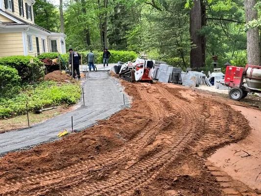 Workers install a paver walkway next to a house, with heavy equipment and dirt tracks nearby on a residential property.