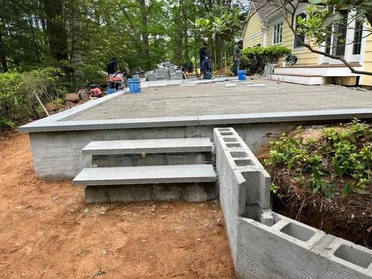 Construction workers build a concrete patio with steps leading to a backyard house exterior.