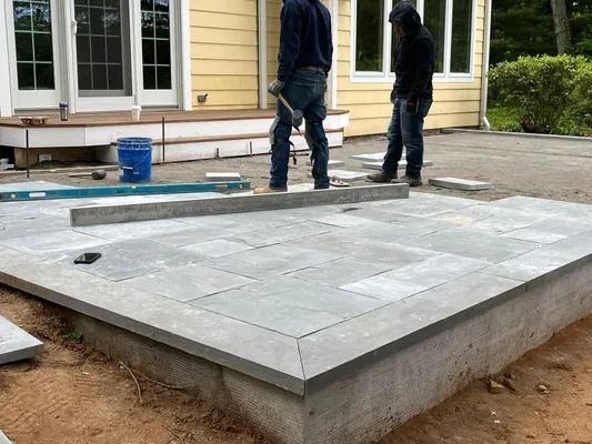 Two people installing stone patio pavers in front of a yellow house.