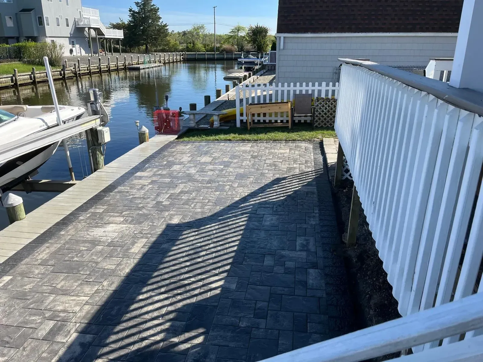 A gray paver patio sits beside a canal with a white railing, a wooden bench, and a boat docked in the background.