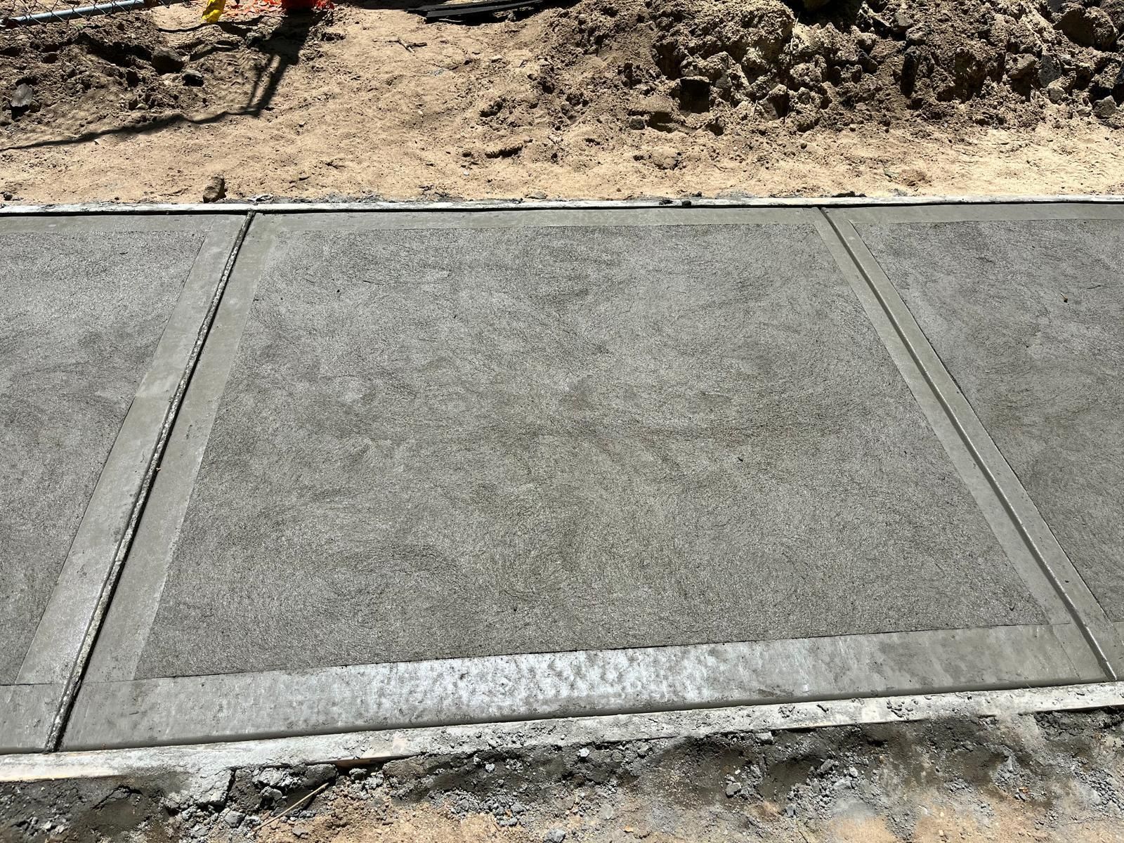 A freshly poured, textured gray concrete sidewalk panel with smooth, troweled borders, adjacent to an earthen trench.
