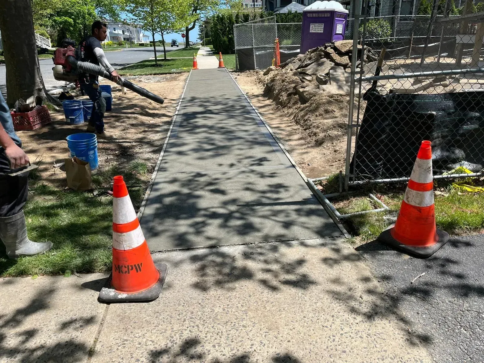 A worker uses a leaf blower on a freshly poured concrete sidewalk marked by traffic cones near a construction site.