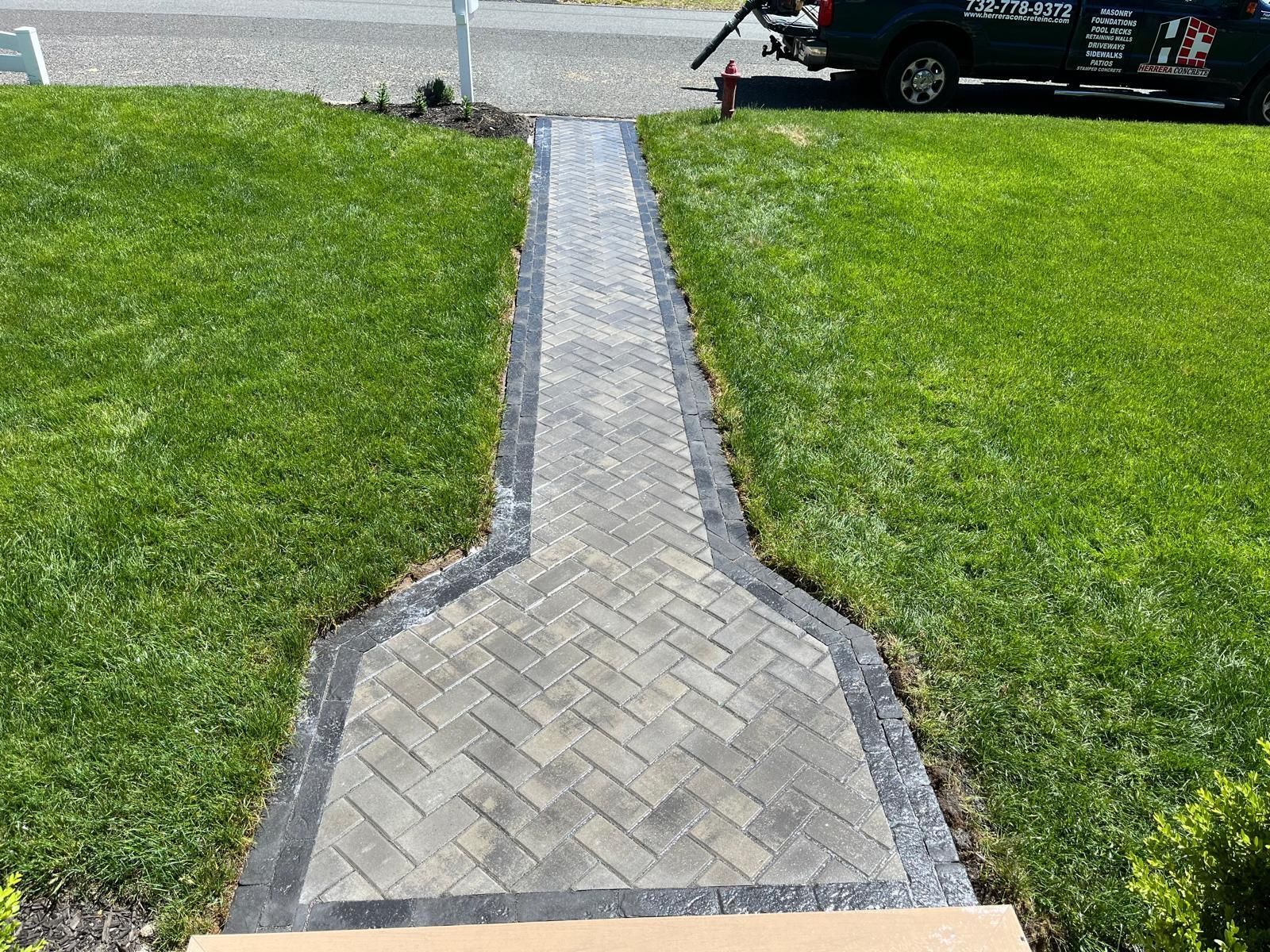 A grey herringbone-patterned brick walkway with a dark border, set between two patches of green lawn leading to a road.