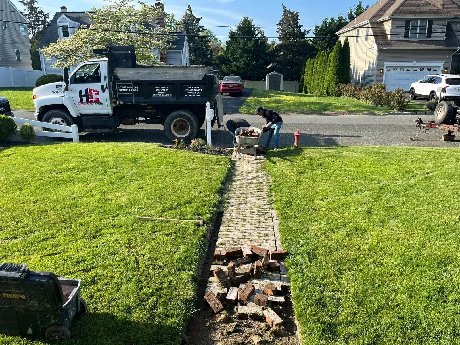 A worker in a yard repairs a brick walkway, with a black dump truck parked on the street in a suburban neighborhood.