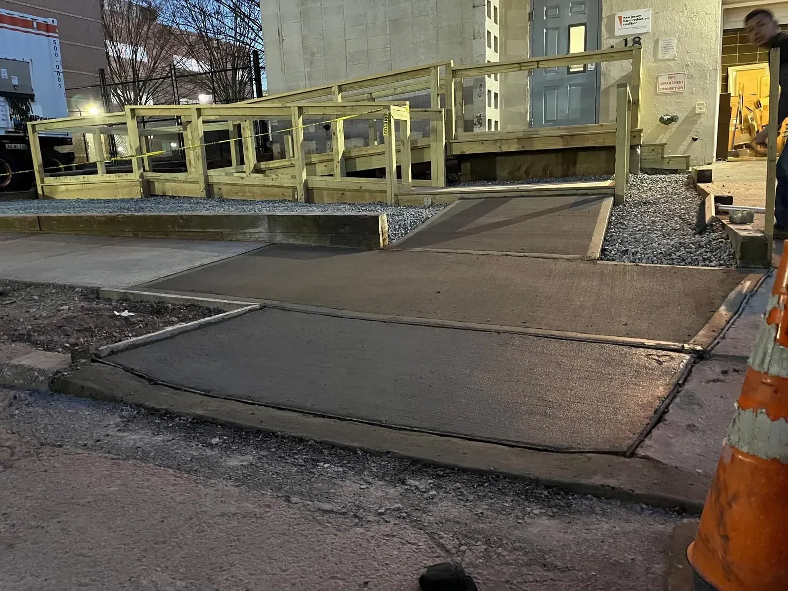 A wooden wheelchair ramp leads up to a building entrance, featuring a freshly poured concrete path at the base.