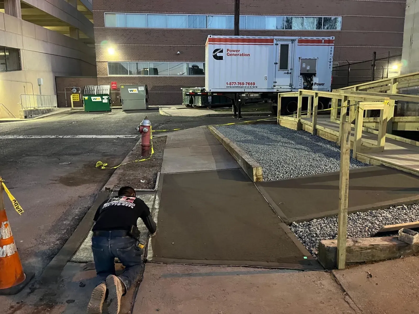 A worker kneels to smooth fresh concrete on a sidewalk path near a temporary generator and a ramp at night.