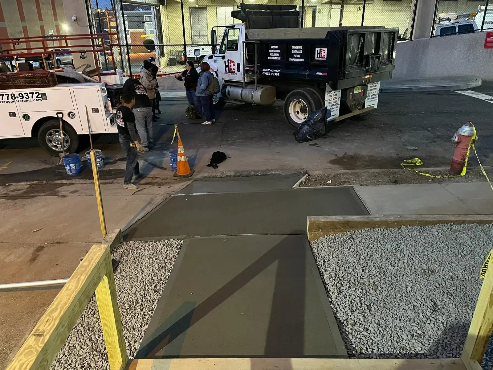 Workers stand near a construction truck next to a freshly poured concrete walkway in a gravel area.