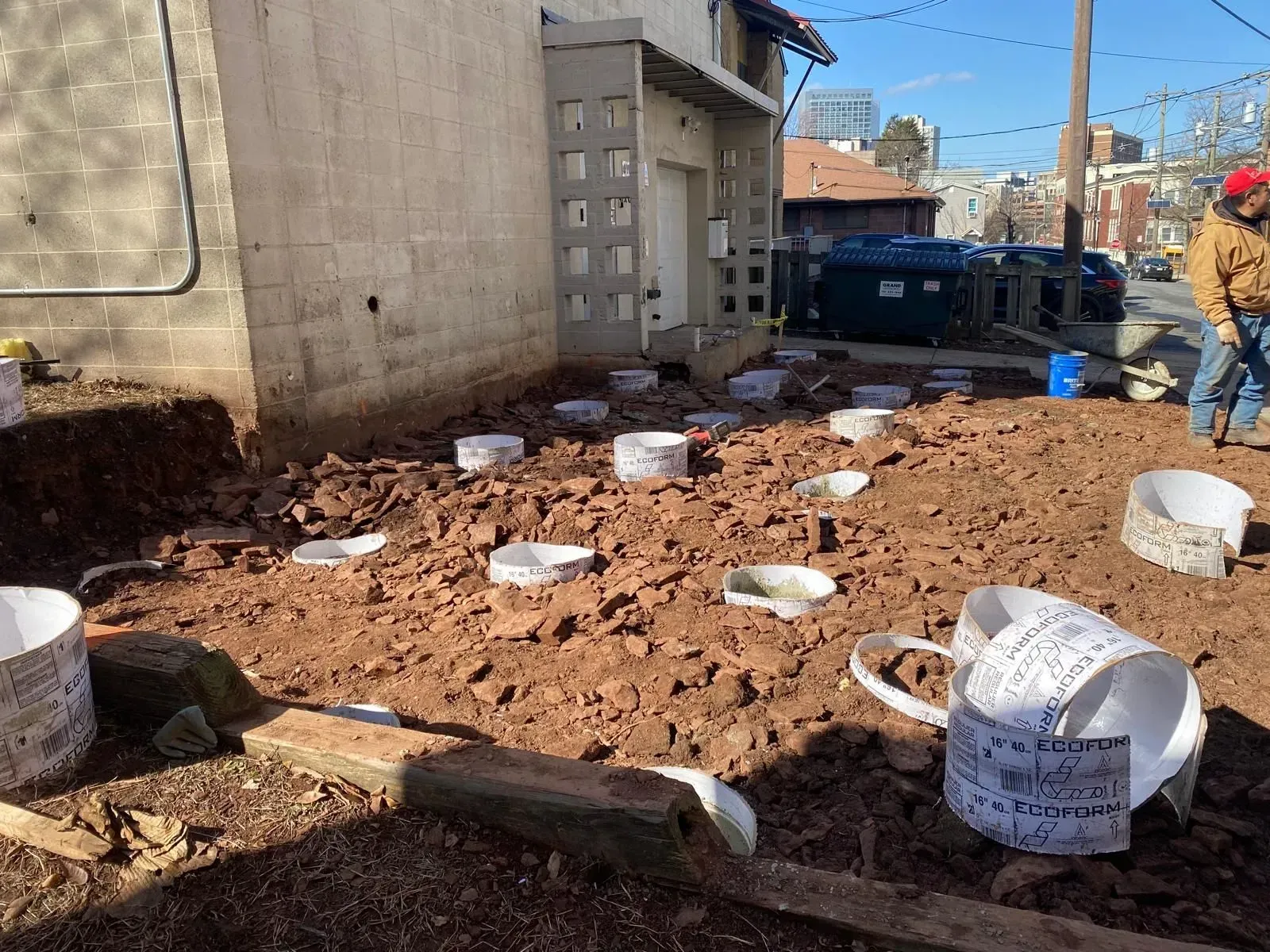 Construction site featuring multiple concrete foundation forms placed in a dirt lot alongside a building.