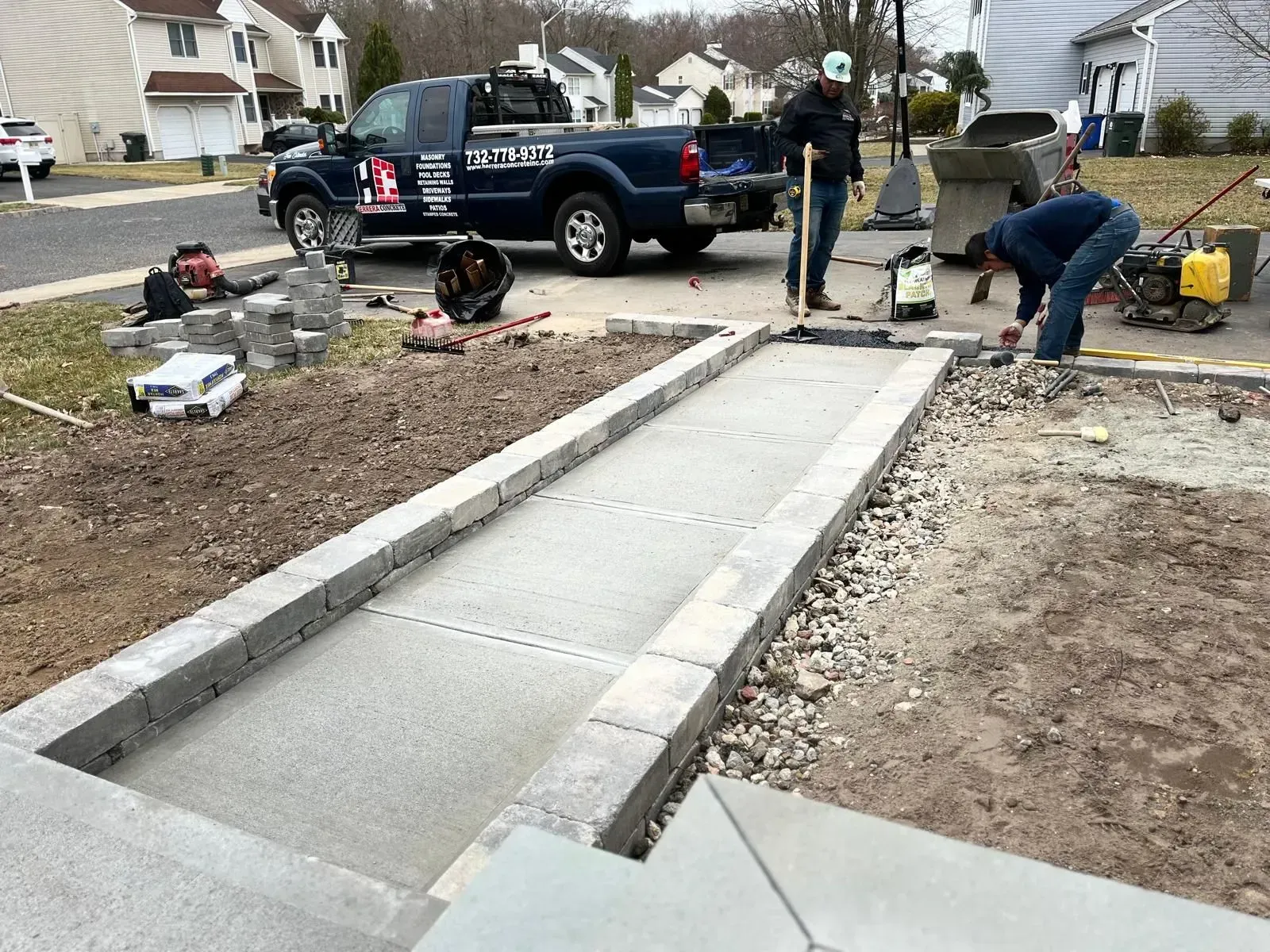 Workers in a suburban driveway construct a concrete path bordered by gray stone blocks near a parked utility truck.