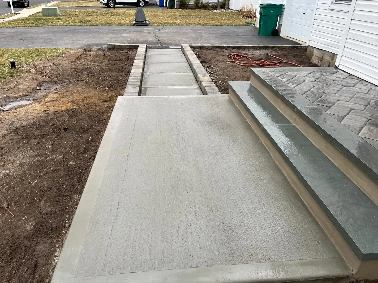 A new concrete walkway leads to a small landing with two steps made of gray, rectangular pavers leading to a house.