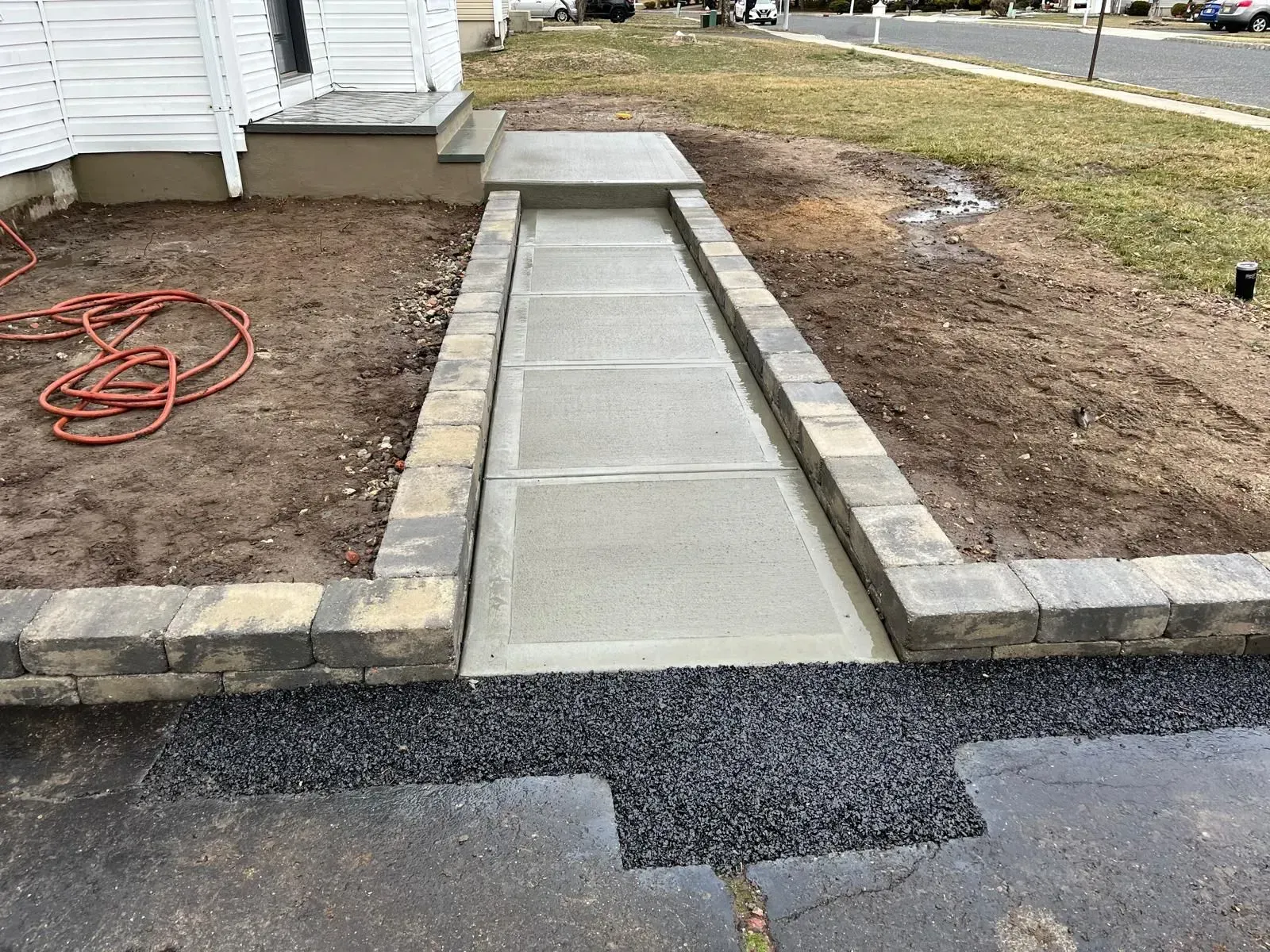 A concrete walkway leads to a house porch, framed by low stone retaining walls and flanked by dirt and an asphalt driveway.