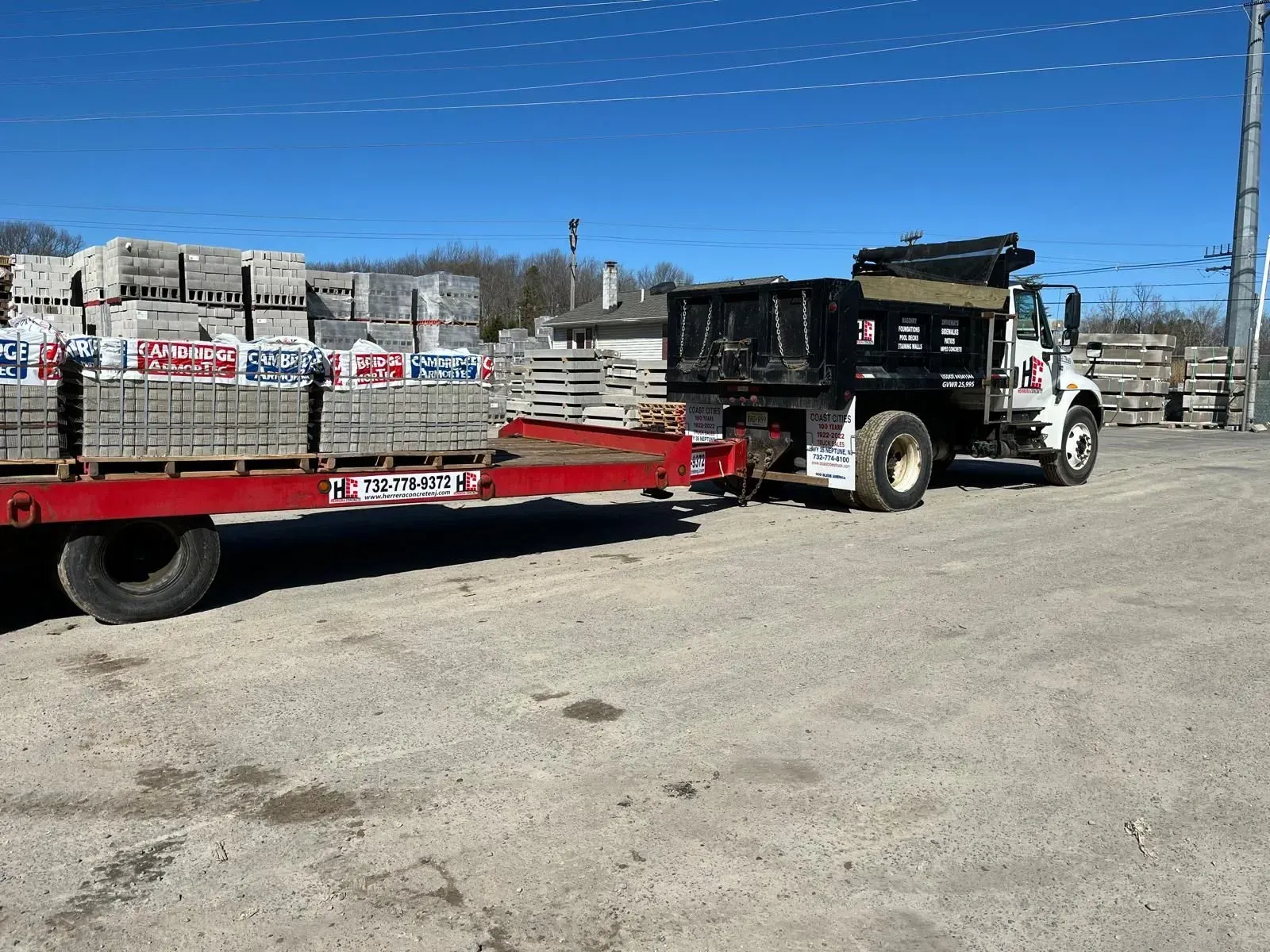 A red flatbed trailer carrying palletized bricks, attached to a black dump truck in a gravel yard under a clear blue sky.