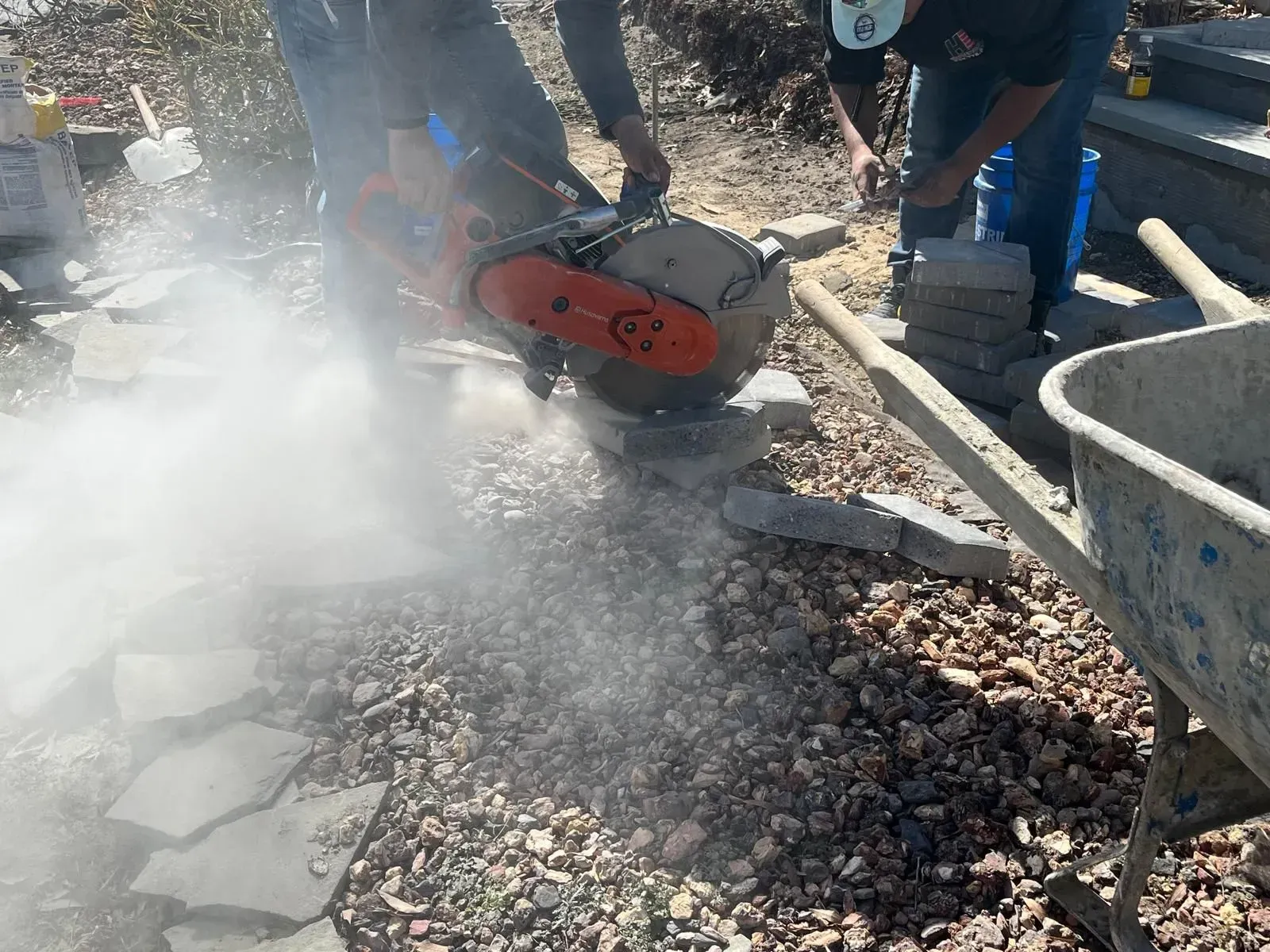 A worker uses an orange gas-powered concrete saw to cut a stone paver, creating a large cloud of dust outdoors.