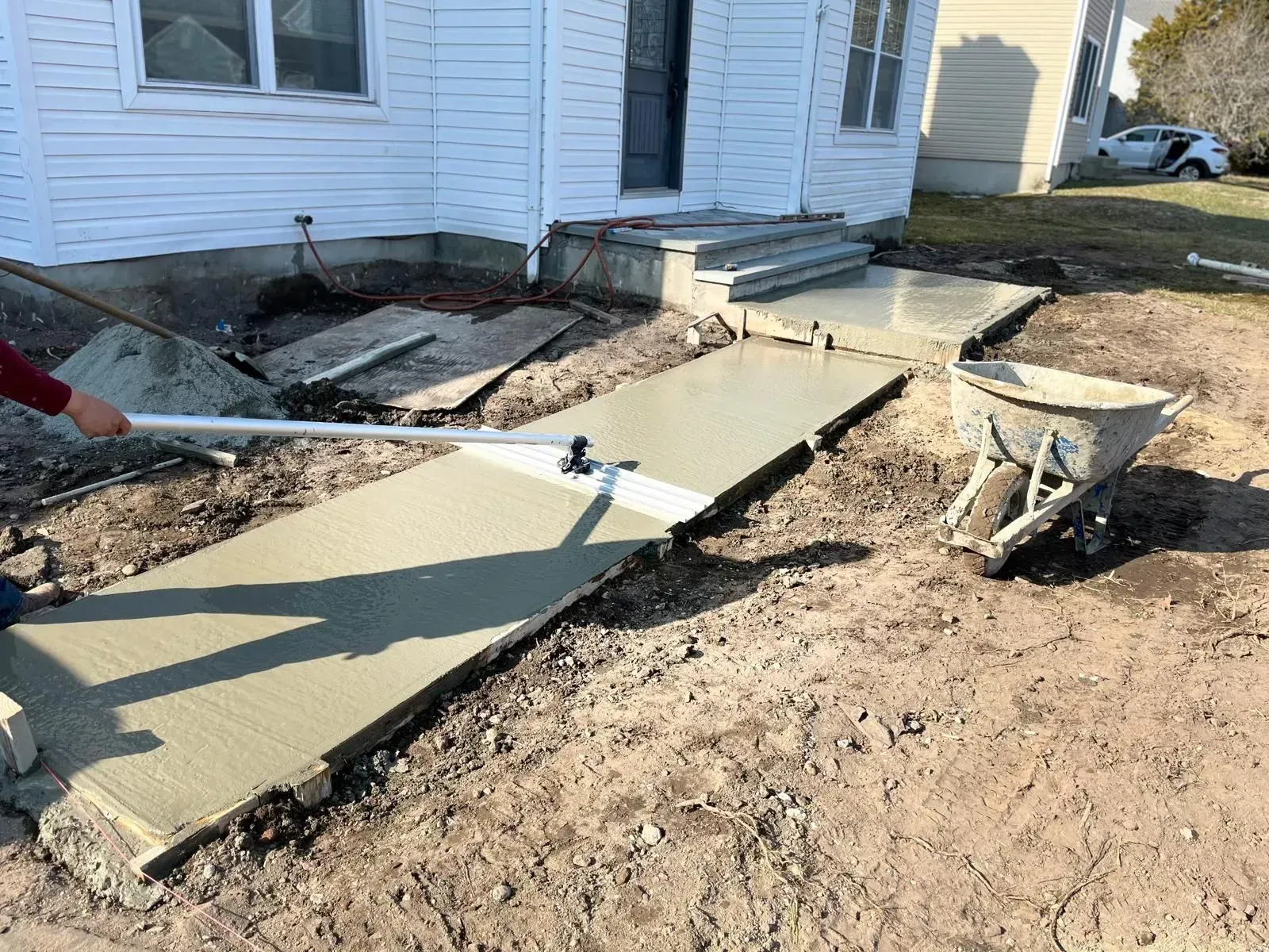 A person uses a long-handled concrete bull float to smooth a newly poured sidewalk leading to a house entrance.