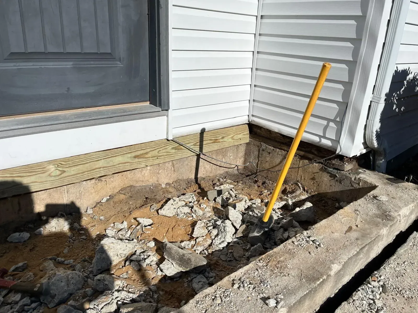 A partially demolished concrete stoop with debris in front of a white house with a dark gray door and a yellow shovel.