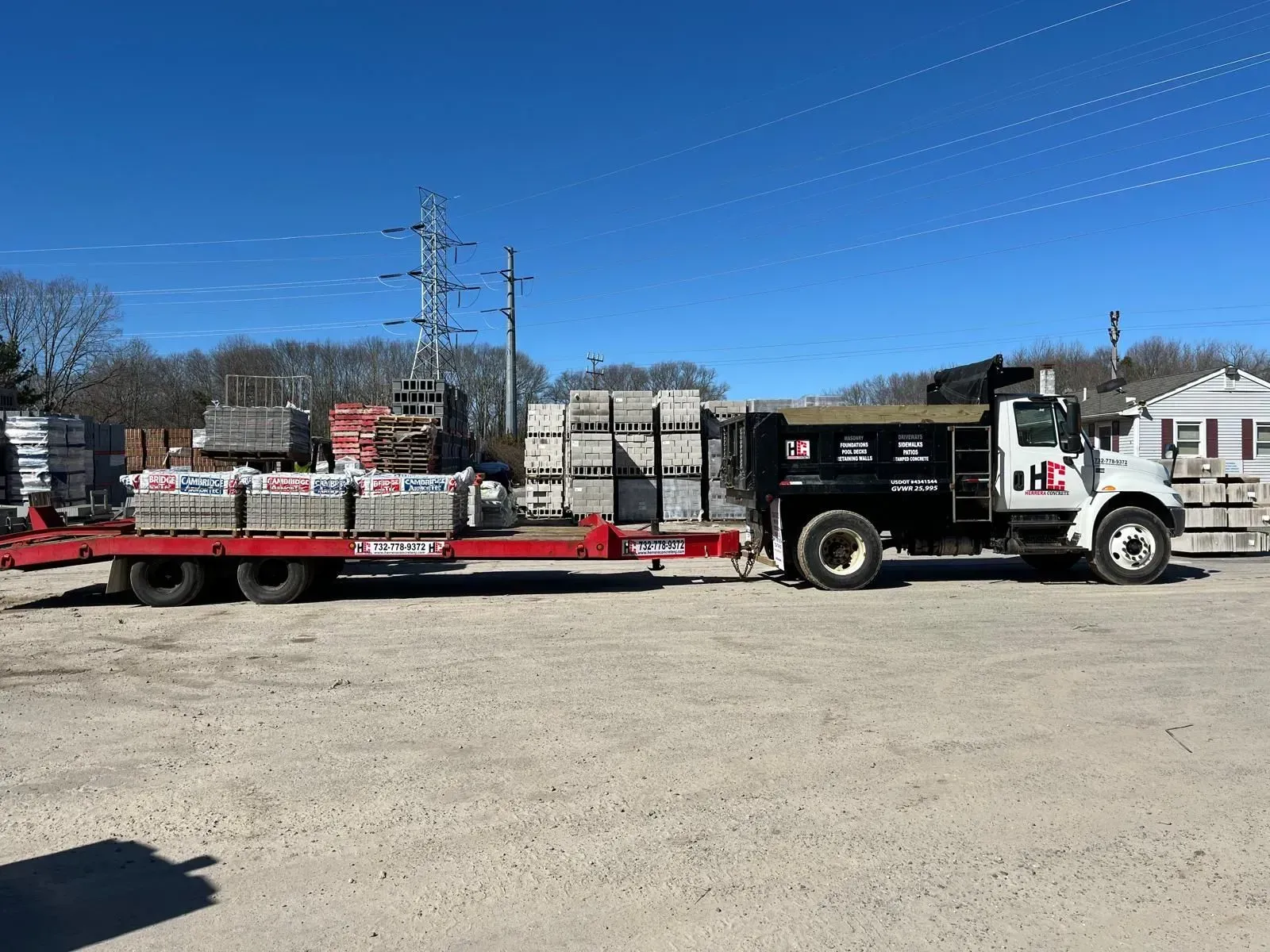 A flatbed trailer carrying stacked masonry blocks is hitched to a black work truck in a gravel yard under a clear blue sky.
