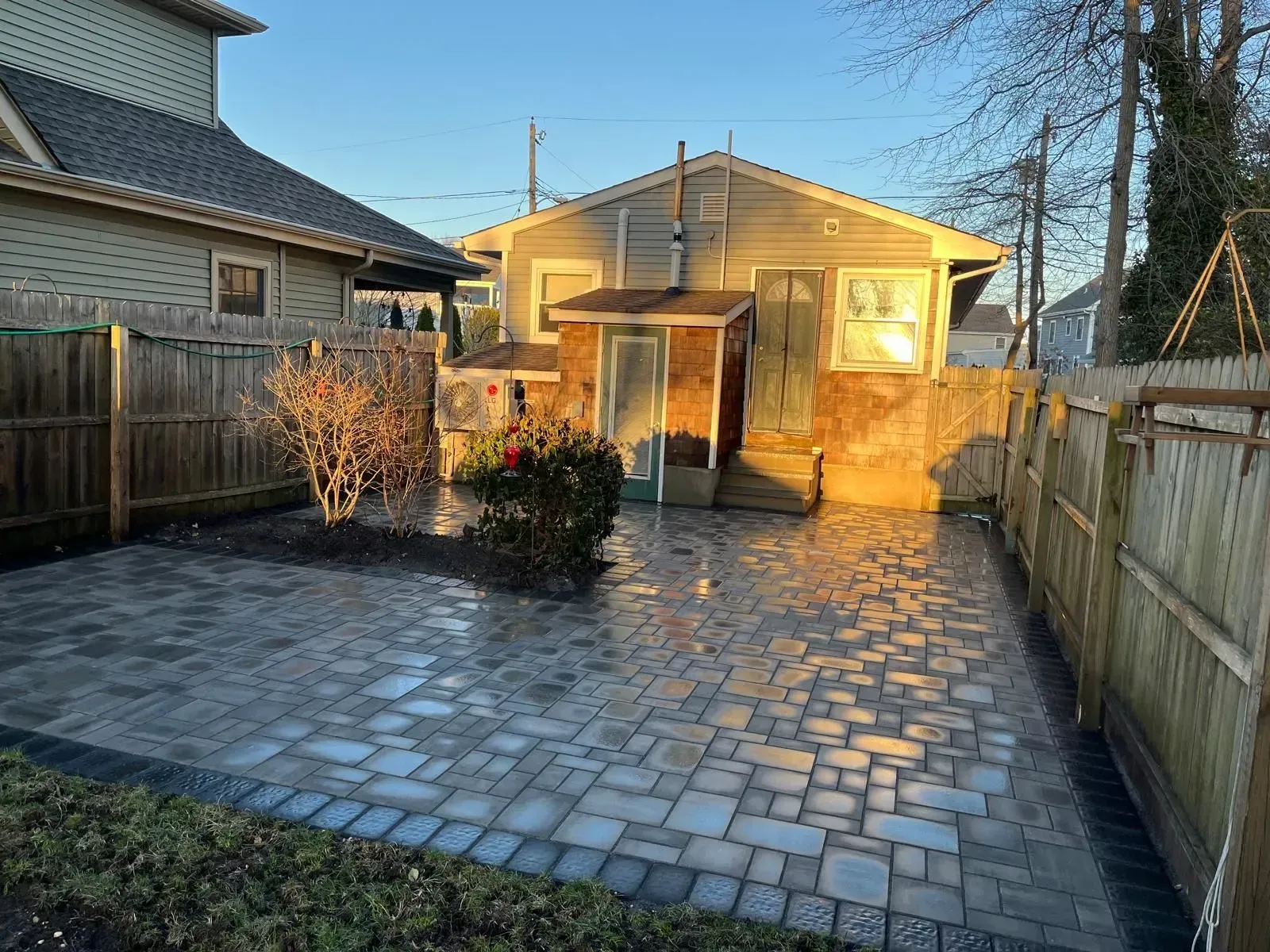 A backyard with a newly installed stone paver patio, a small shrub, and wooden fencing, viewed in warm sunset light.