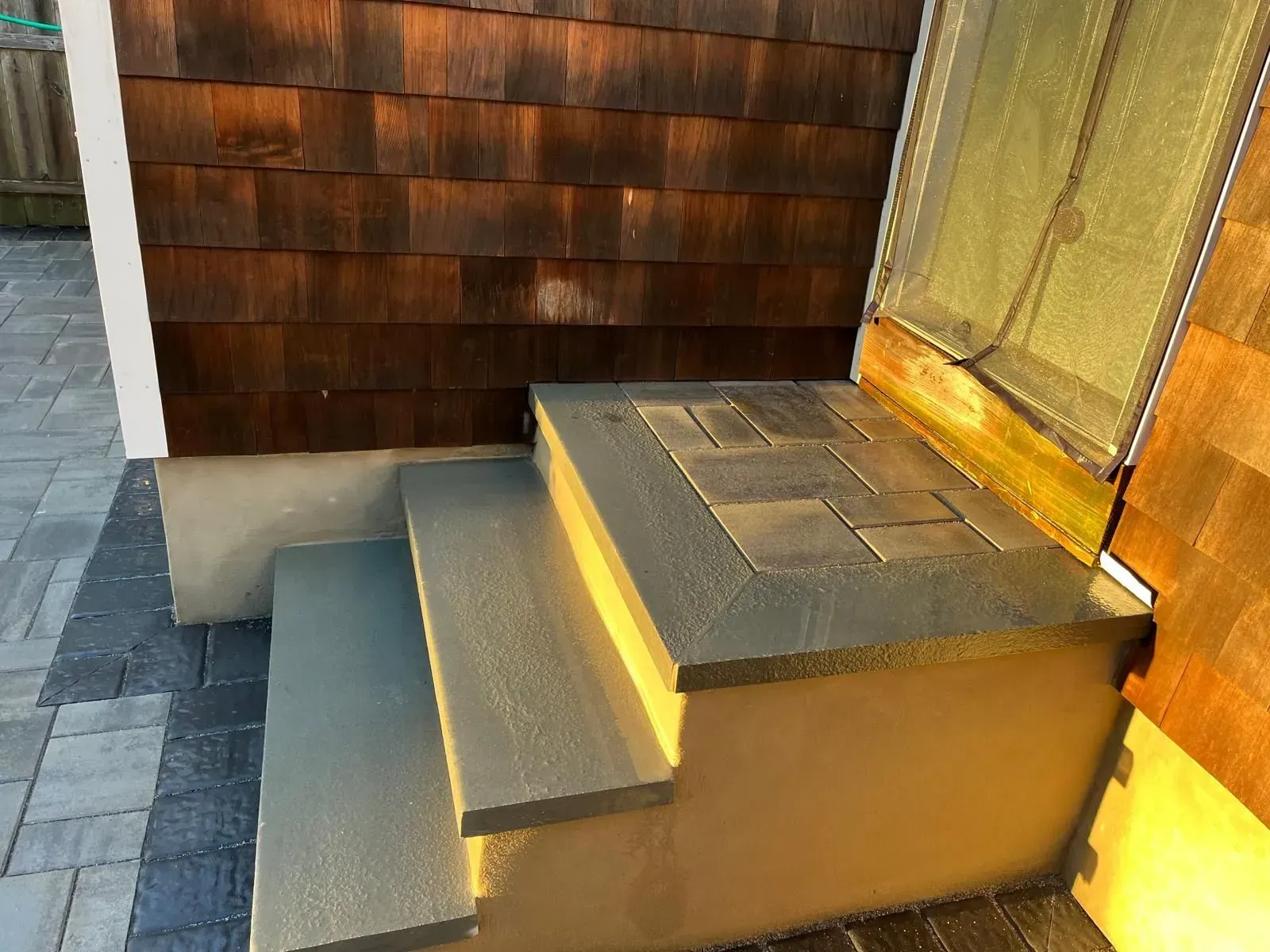 Two concrete steps lead up to a doorway with a screen door, set against a house with dark brown wood shingle siding.
