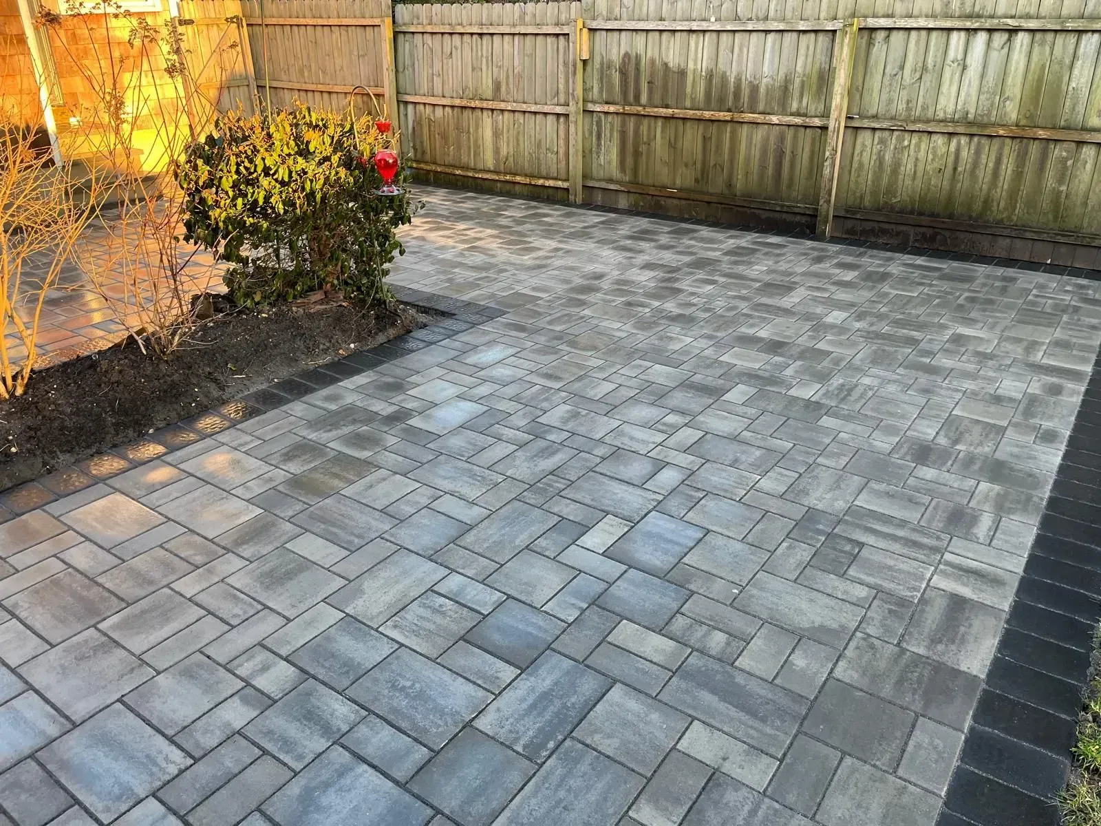 A patio paved with rectangular gray stone bricks in a basket-weave pattern, bordered by dark charcoal bricks near a fence.