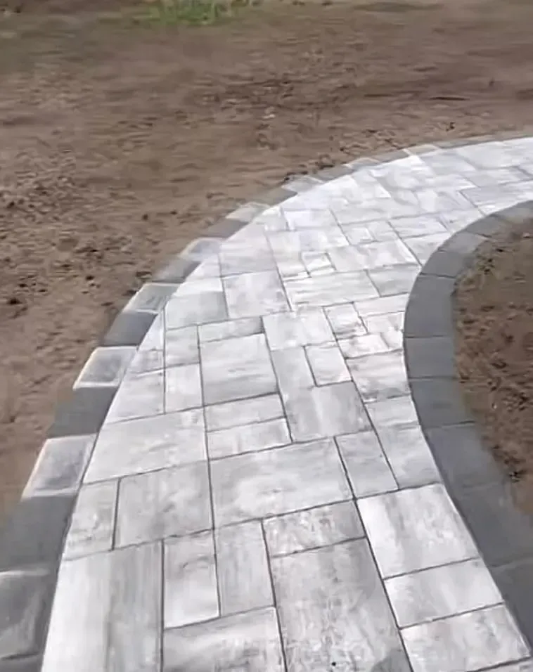 A curved walkway made of light gray rectangular pavers bordered by dark gray stones, set against bare dirt.