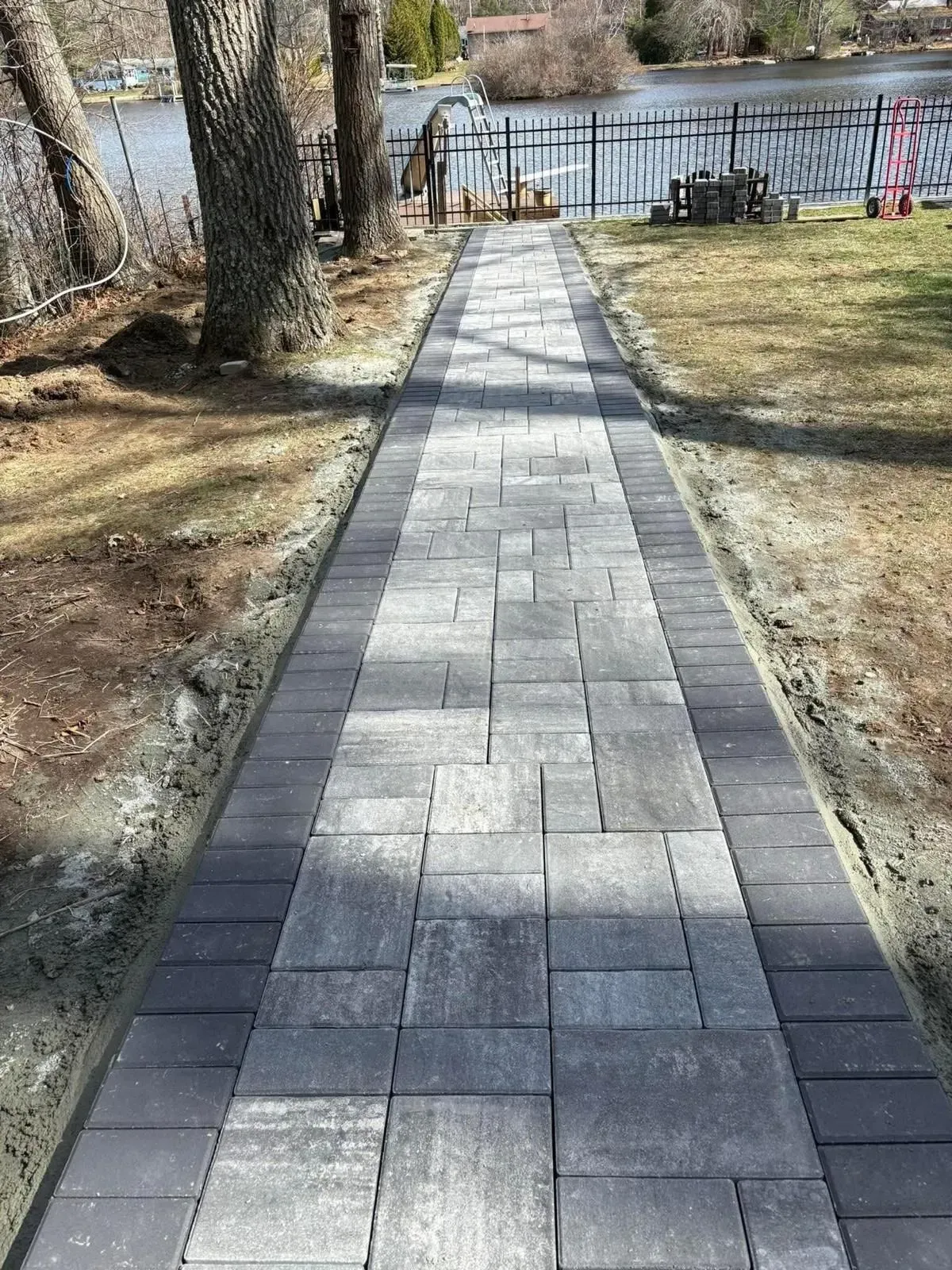 A gray stone paver walkway with dark border stones leads toward a waterfront property with trees and a black metal fence.