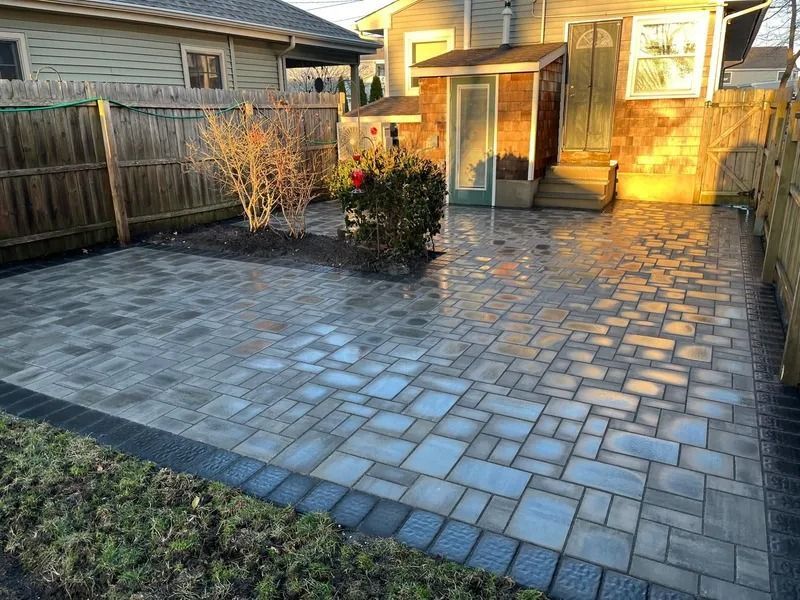 A backyard patio made of grey rectangular pavers with a dark border, next to a house and wooden fence at sunset.