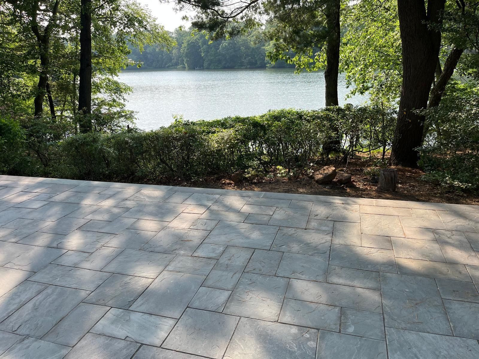 A gray stone patio overlooks a calm lake framed by lush green trees and shrubs.