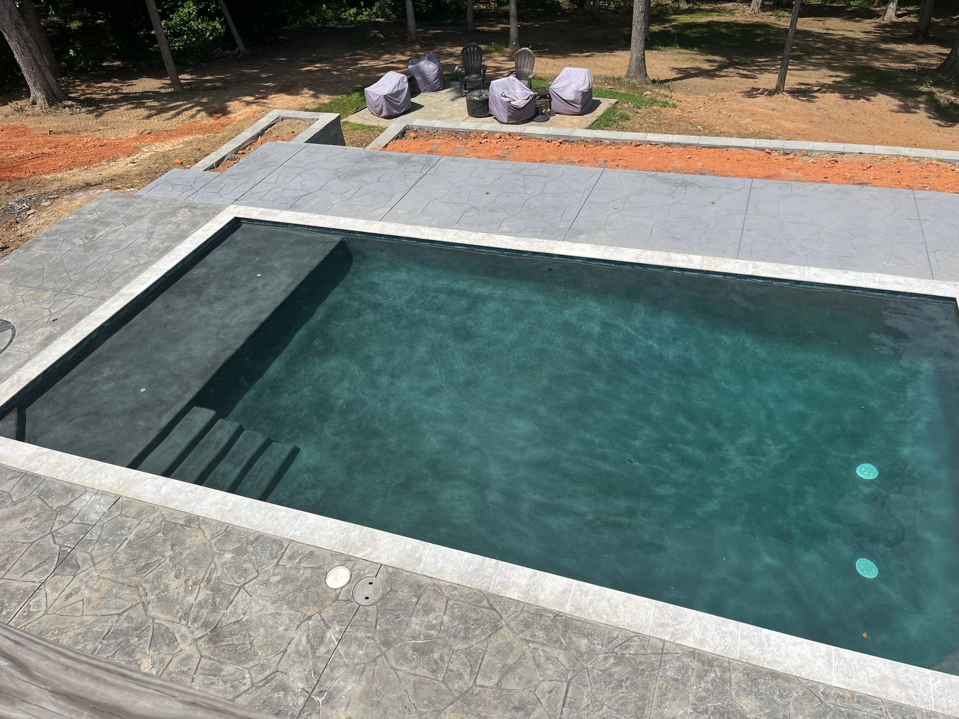 An elevated rectangular swimming pool with steps, surrounded by gray concrete. Lawn and trees in the background.