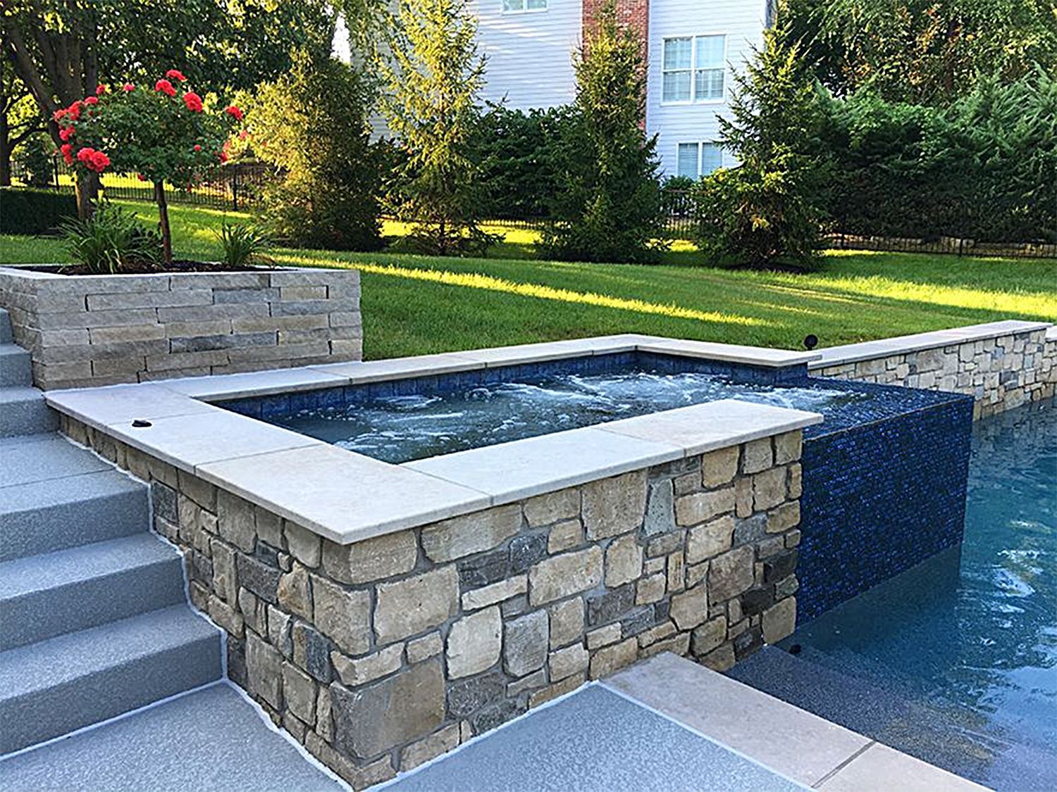 Stone-walled hot tub next to a pool, with steps and grassy lawn in the background.