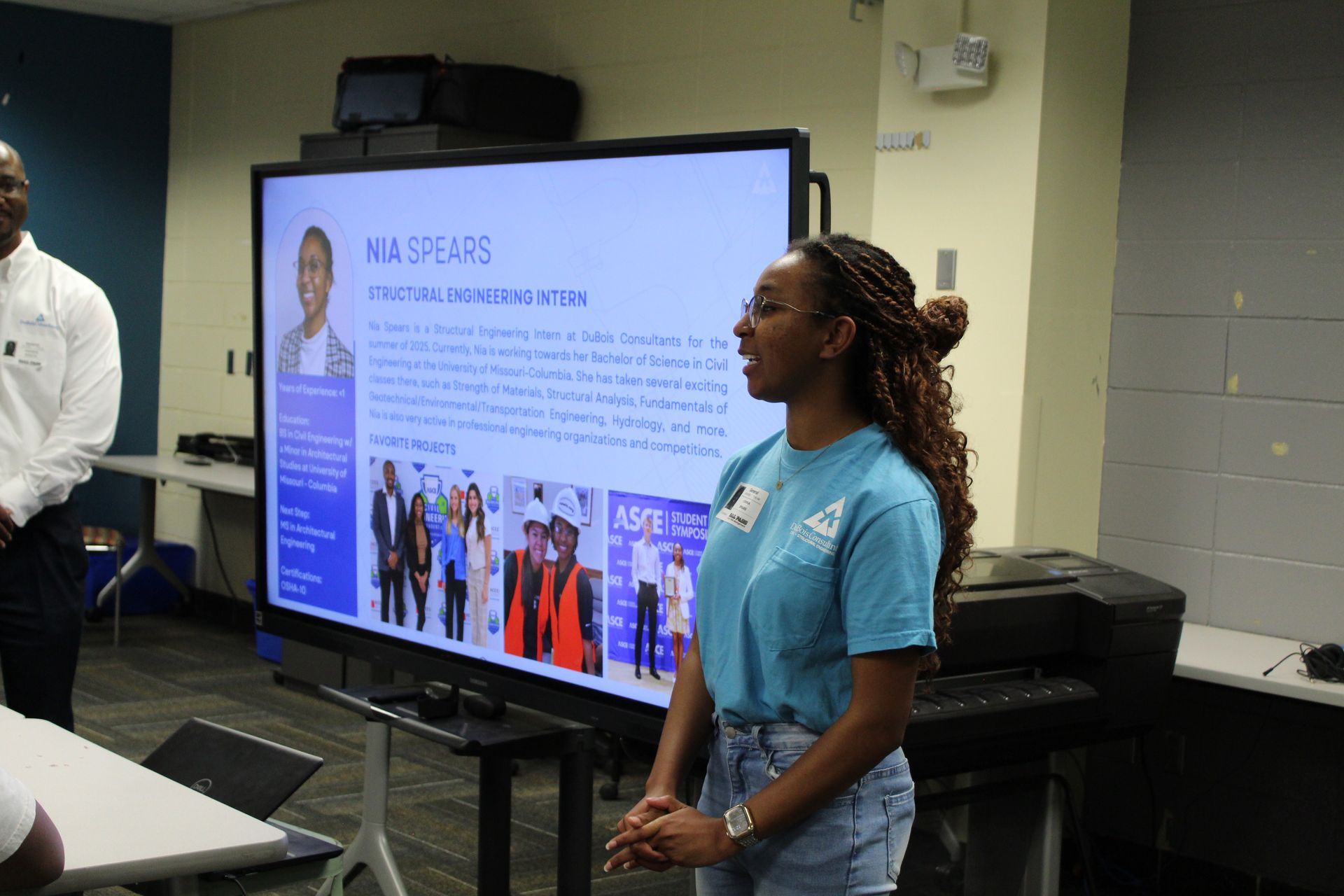 Woman presenting at a screen; man stands to the left.  Presentation about 