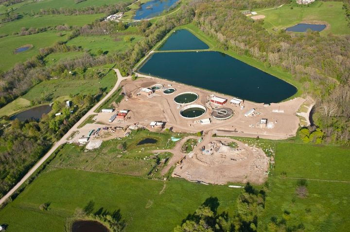 Aerial view of a wastewater treatment facility with circular tanks beside a dark green pond and fields.