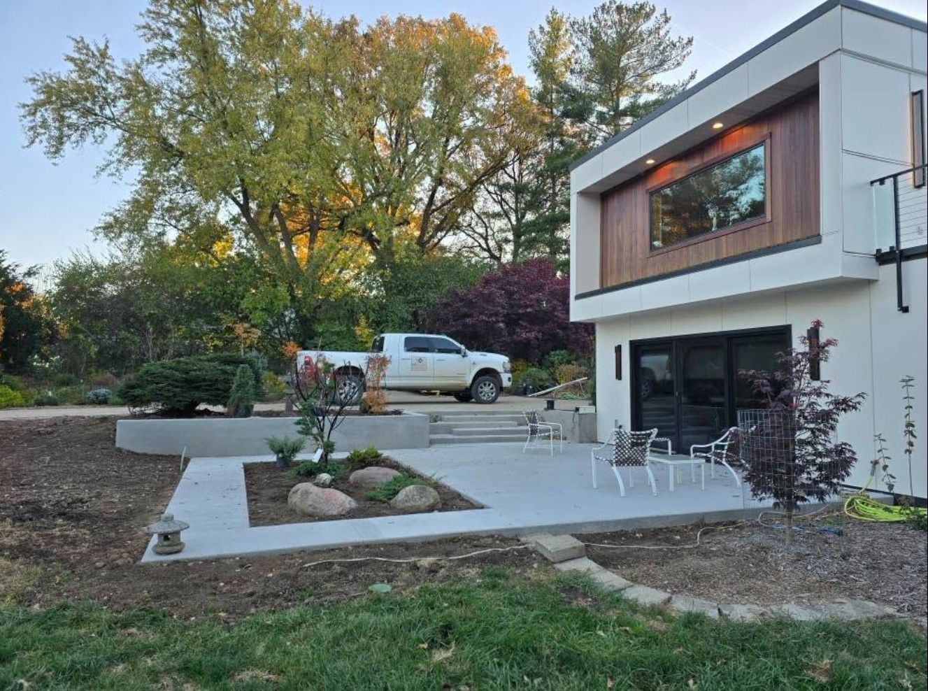 A modern home with a white exterior, wood accent, and patio overlooks a landscaped garden with a parked white pickup truck.