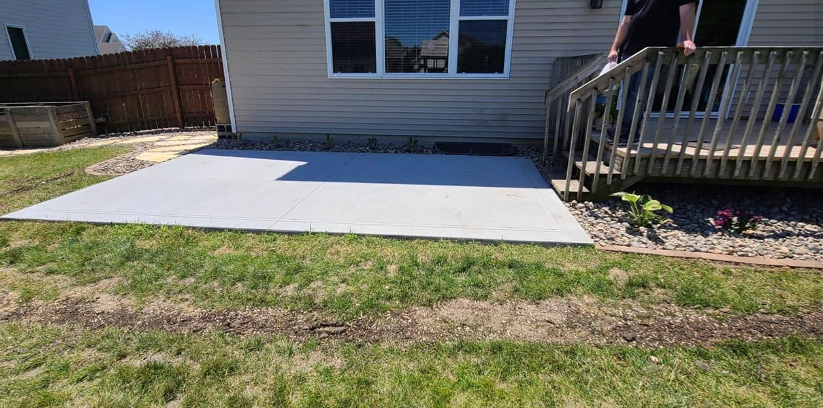 A newly poured concrete patio sits in a backyard next to a house with beige siding and a wooden deck.