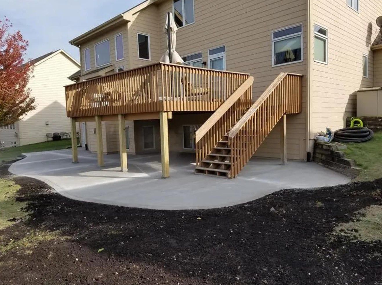 A wooden deck with stairs attached to a beige house, overlooking a newly poured concrete patio in a backyard setting.
