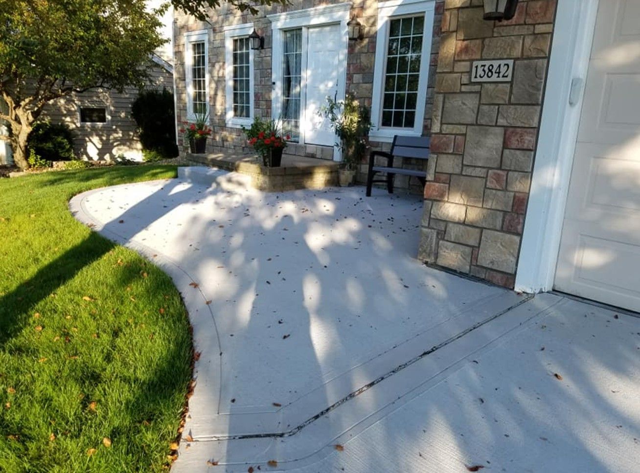 A concrete driveway or patio curves alongside a suburban home with stone siding, a house number, and a small bench.