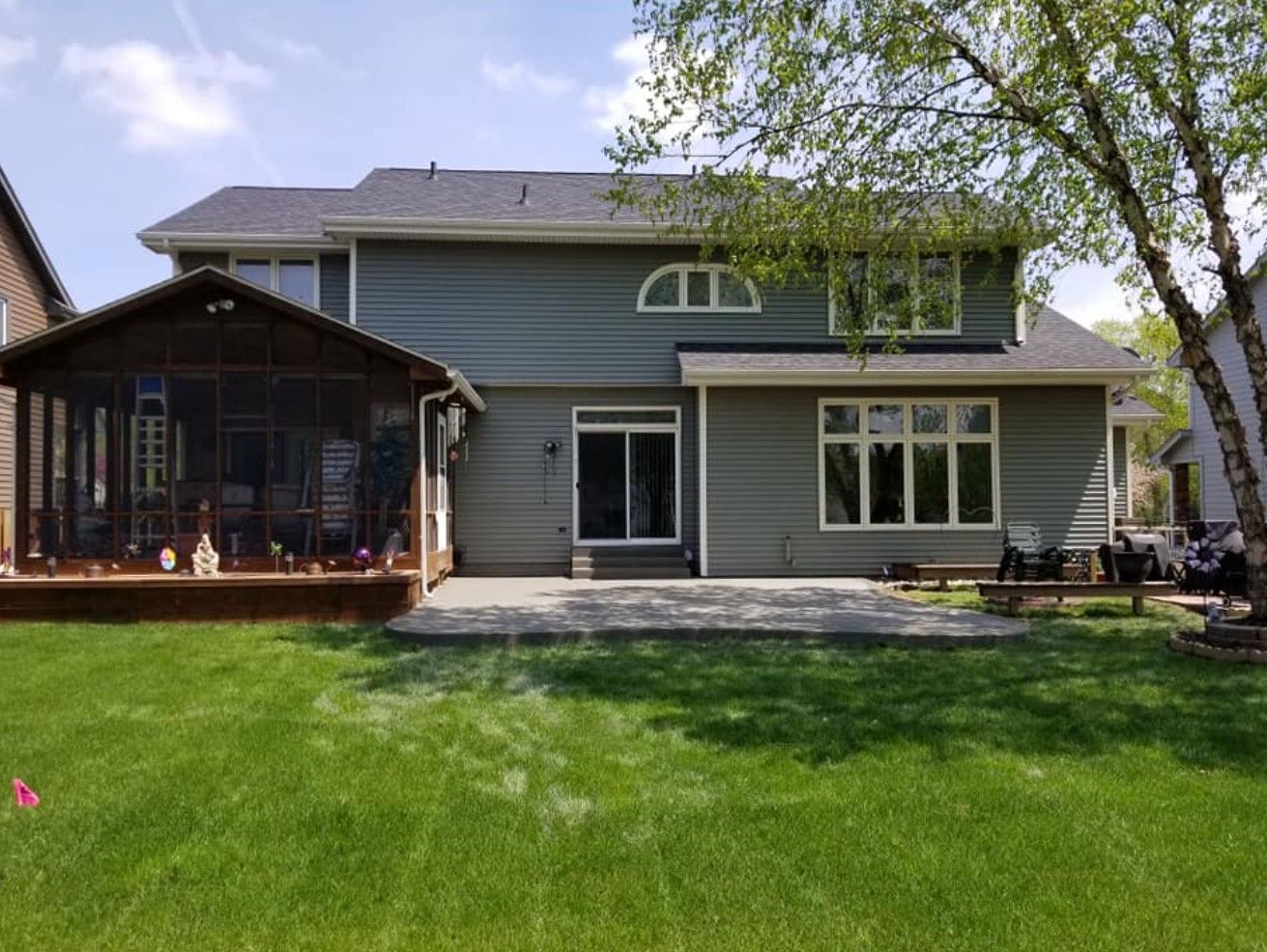 A two-story grey house with a wooden screened-in porch, a concrete patio, and a large green backyard on a sunny day.