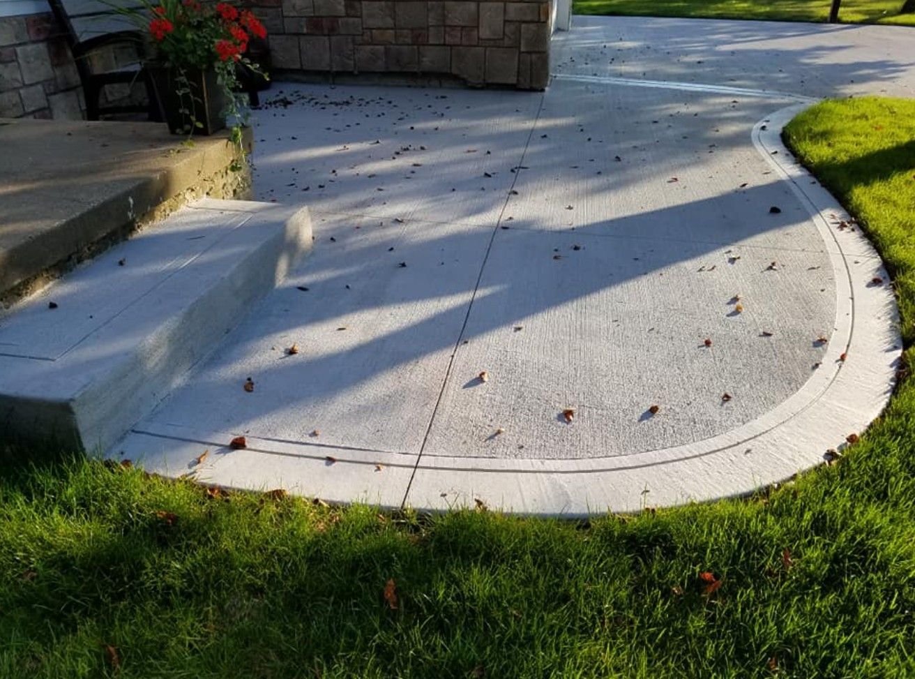 A light gray concrete patio with a curved stone border edge, next to a grass lawn and stone steps.