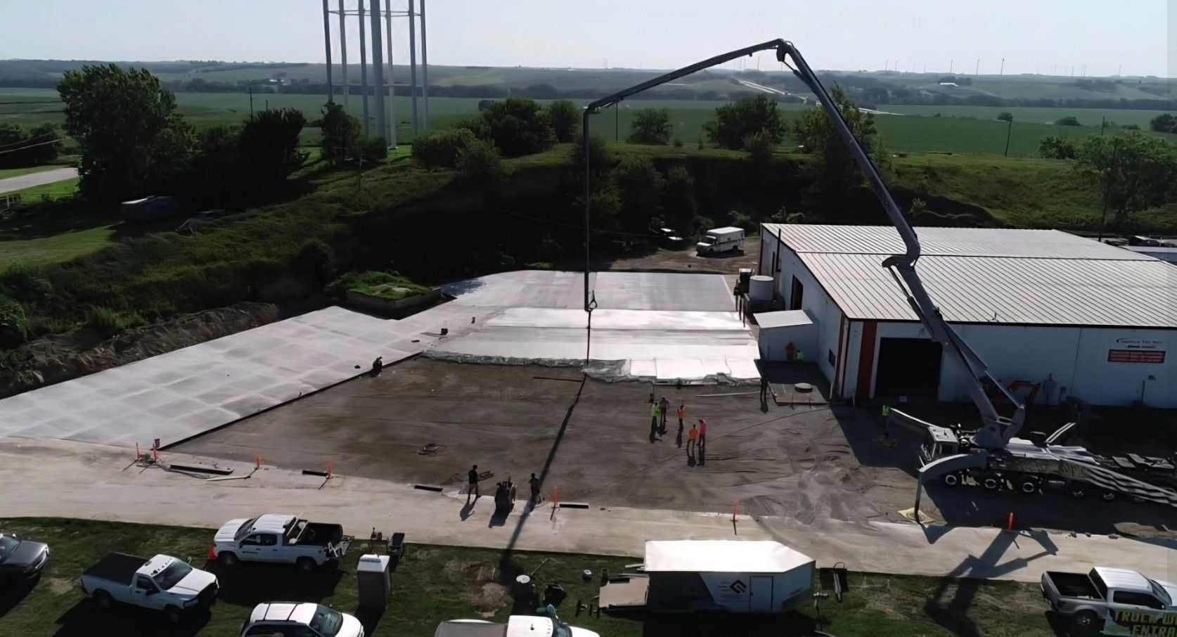 An aerial view of a construction crew pouring concrete for a large outdoor slab next to a white warehouse building.