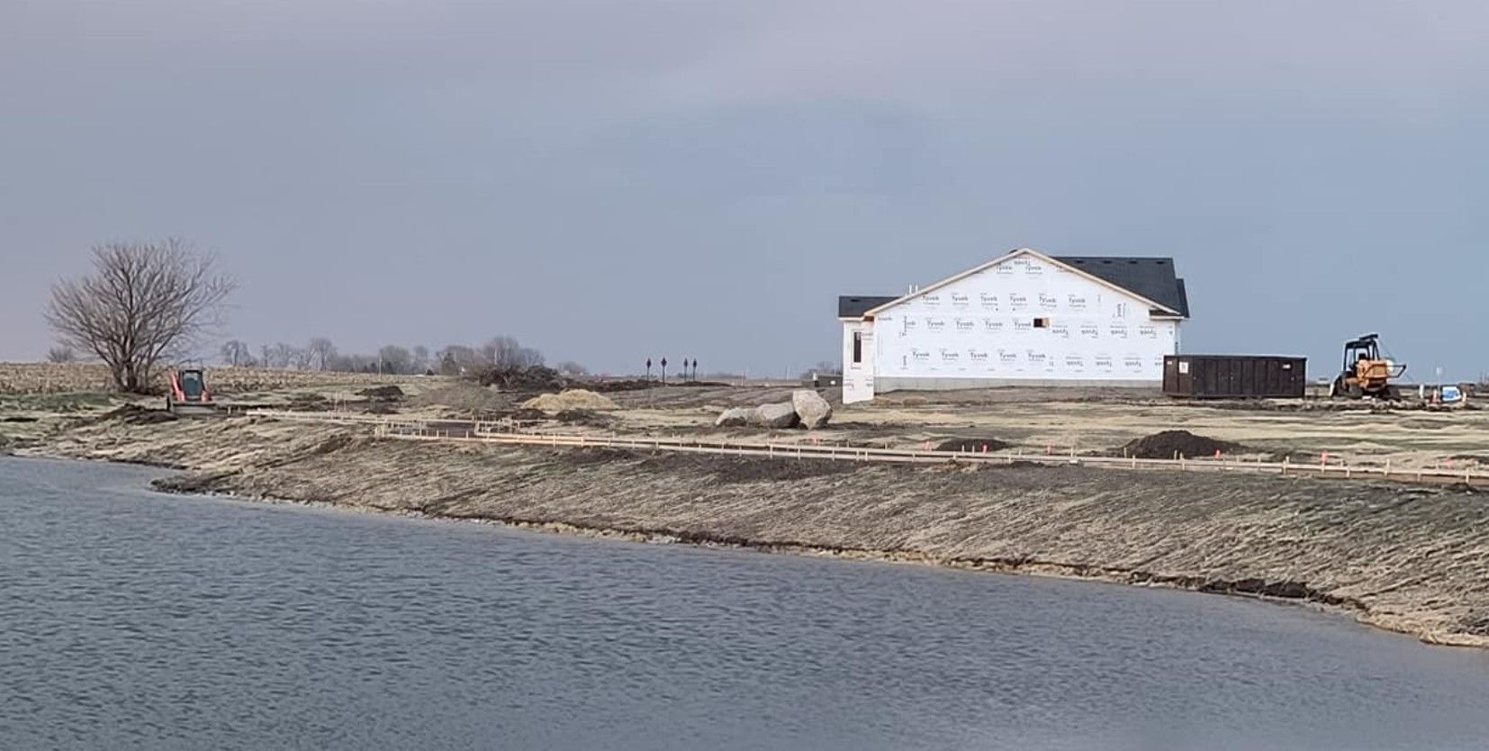 A white house under construction stands on a barren, dirt-covered lakeshore under a gray, overcast sky.